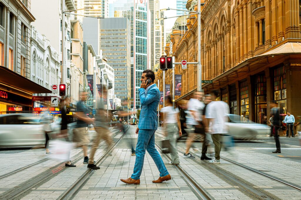 A businessman in a blue suit crosses a busy street in Sydney, Australia, surrounded by motion-blurred pedestrians.